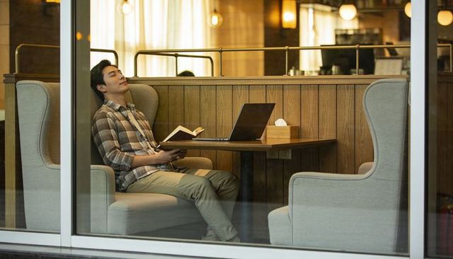 Asian man resting in cozy cafe booth reading book with laptop, warm relaxed mood