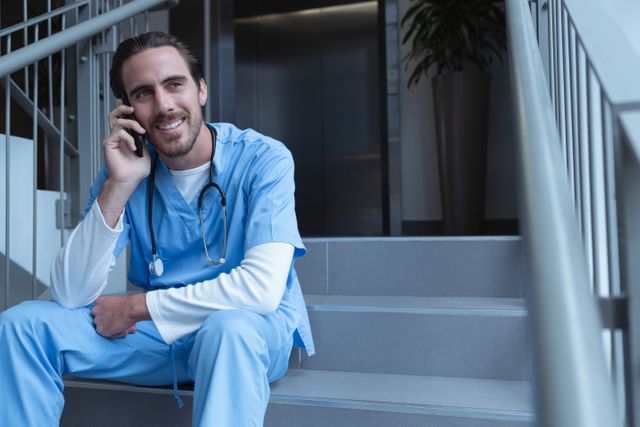 Front view of happy male surgeon talking on mobile phone while sitting on stairs at hospital