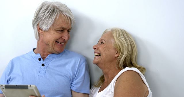 A senior Caucasian couple shares a joyful moment together while looking at a tablet, with copy space. Their laughter and close interaction suggest a warm, loving relationship.