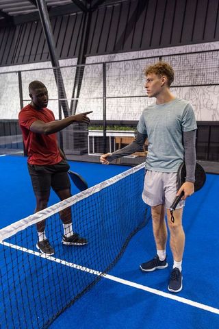 Two male players gesturing and readying serve on vibrant blue indoor padel court with rackets