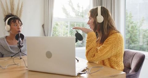 Two women recording podcast at home using laptop, microphones and headphones