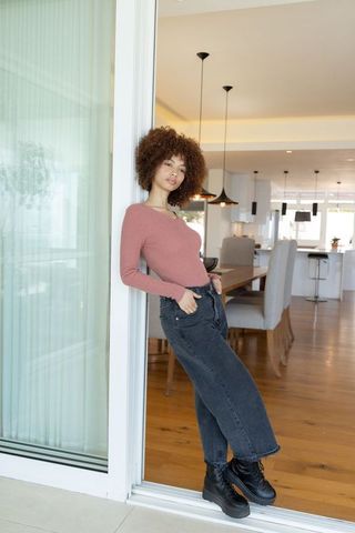 African american woman leaning in glass doorway at modern open-plan home wearing dusty-rose top