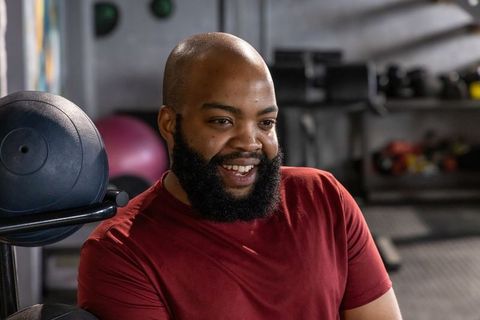 Smiling african american man resting on gym bench wearing red shirt during workout