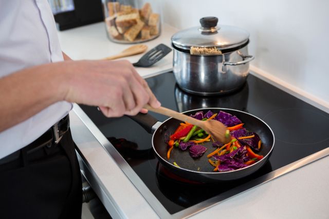 Man cooking colorful vegetables in a frying pan on a modern stove in a home kitchen. Ideal for use in articles or advertisements related to healthy eating, home cooking, culinary skills, and domestic lifestyle.