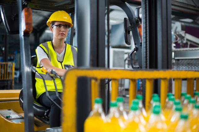 Female factory worker driving forklift in an industrial warehouse, wearing safety gear including a hard hat and high visibility vest. Ideal for use in articles or advertisements related to industrial work, manufacturing, logistics, workplace safety, and women in industry.
