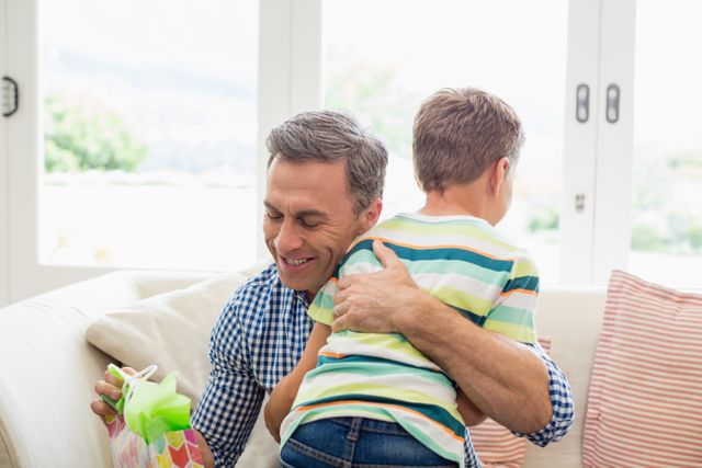 Father and son sharing a heartfelt embrace in a cozy living room. The father holds a gift, suggesting a special occasion or celebration. Perfect for use in family-oriented advertisements, parenting blogs, or articles about family relationships and bonding.