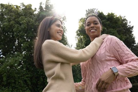 Smiling diverse women celebrating outdoors in sunlit park wearing tiaras and cozy sweaters