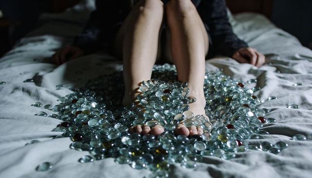 Woman sitting on bed with feet covered in glass pebbles, moody low-key intimate scene
