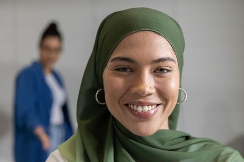Smiling woman wearing green headscarf and hoop earrings with coworker blurred in office