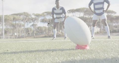 White rugby ball on pink kicking tee with players lining up for kickoff on grass pitch