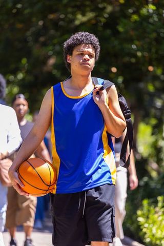 Young african male walking with basketball in blue yellow jersey sunlit park with backpack
