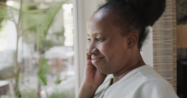 Happy senior african american woman with hand on face looking out of window at home. Retirement, domestic life, solitude and senior lifestyle, unaltered.