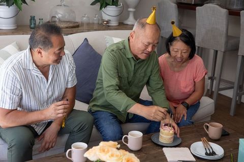 Asian family celebrating birthday at home cutting cake on sofa wearing party hats cozy celebration