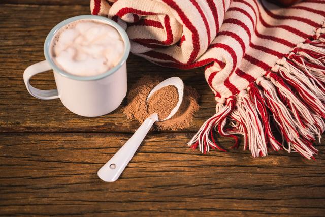 Warm and inviting scene featuring a mug of hot chocolate with frothy milk, a spoonful of cocoa powder, and a red and white striped blanket on a rustic wooden table. Ideal for use in lifestyle blogs, winter-themed promotions, home decor advertisements, and social media posts about comfort and relaxation.
