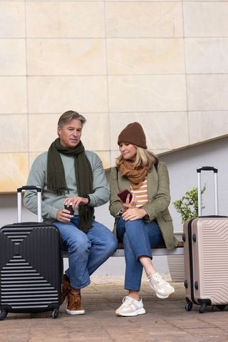 Mature couple waiting with luggage on bench at transport hub holding coffee and checking phone
