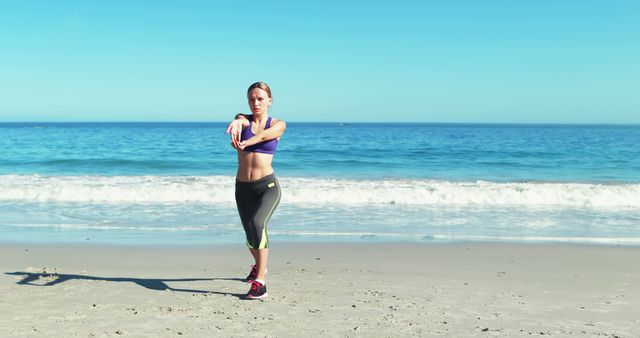 Woman stretching arms on beach during morning workout, wearing sportswear, with ocean and waves in background. Useful for content related to fitness, outdoor exercise, healthy lifestyle, summer activities, and yoga sessions.