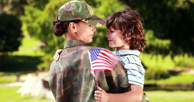 Shows a mother in camouflage military uniform holding her young son while he waves an American flag. Captures strong sense of patriotism and familial love. Can be used for advertisements celebrating military families, promotional material supporting the troops, and campaigns around national holidays such as Memorial Day, Veterans Day, and the Fourth of July.