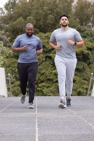 Two men jogging together on tree-lined paved park path wearing sportswear and smartwatches