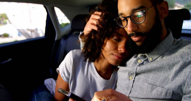 Couple sitting close together in back seat of a car, embraced affectionately, one holding a mobile phone. Ideal for themes of love, family, relaxation, travel, and modern lifestyle.
