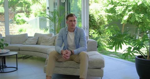 Thoughtful young man sitting in modern living room near sliding doors with garden view
