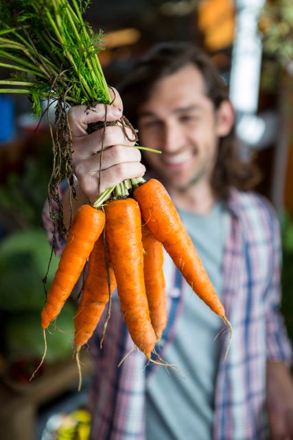 Man holding a bunch of fresh carrots in an organic market. Ideal for promoting healthy eating, local produce, sustainable agriculture, and organic markets. Can be used in advertisements, blogs, and articles related to farming, nutrition, and eco-friendly lifestyles.