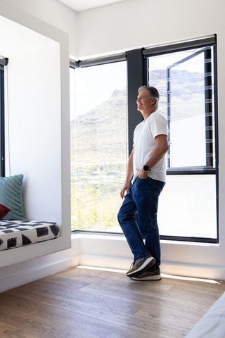 Senior man gazing through large bay windows at sunlit mountain valley, relaxed home moment