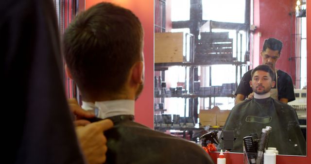 A man gets a haircut in a modern barbershop. The image captures him from behind, with his reflection in the mirror, showing the barber working on his hair. Ideal for use in articles or promotions related to grooming, men’s hair care, barbershop services, and self-care routines.