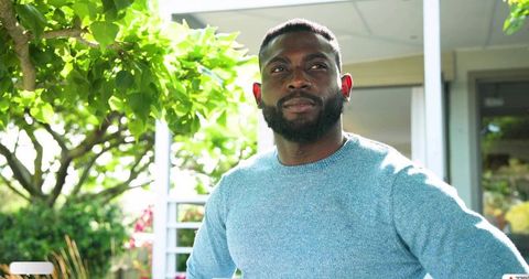 Standing man gazing upward on sunlit porch wearing light blue knit sweater, contemplative