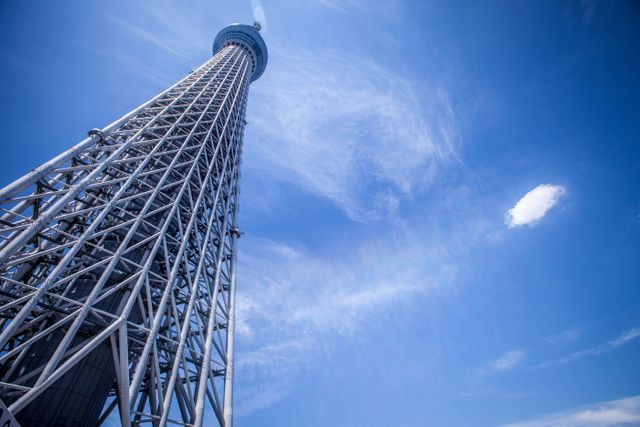 Tokyo Skytree stands tall against a bright blue sky with light clouds, showcasing its impressive steel architecture. Perfect for use in travel brochures, architectural studies, urban landscape features, or promotional materials highlighting modern landmarks and attractions in Japan.