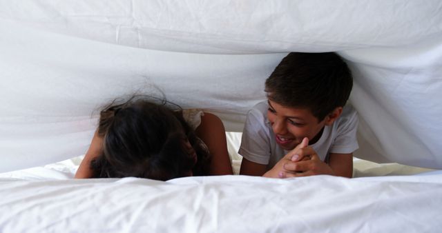Two children, a boy and a girl, enjoying time under a white blanket, embodying sibling bonding and fun. This can be used in parenting blogs, family services promotions, or for illustrating concepts of childhood joy and domestic life.