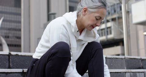 Senior woman tying sneaker laces on city steps in white hoodie and black leggings