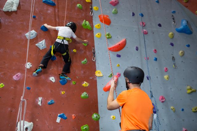 Trainer in orange shirt and helmet assisting boy in safety gear climbing an indoor rock wall. Ideal for promoting fitness, teamwork, youth sports programs, and gym facilities. Highlights the importance of safety in climbing activities.