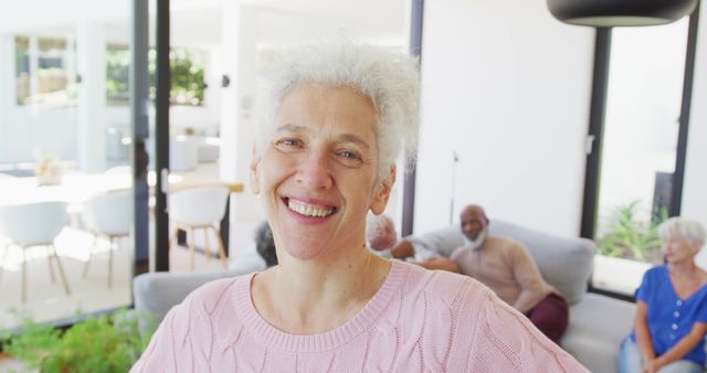 Elderly woman smiling cheerfully in a modern, bright living room with natural light. Other seniors are seen in the background socializing. Useful for themes on aging, senior community, happiness, social connections, living comfortably in old age, and modern home interiors.