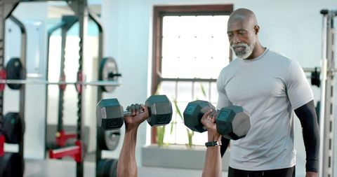 African American Senior Trainer Spotting Client Pressing Dumbbells on Bench in Modern Gym