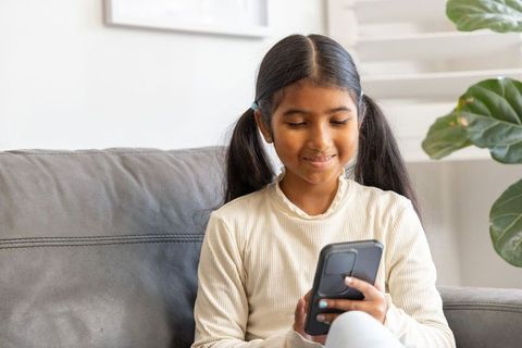 Indian girl sitting on sofa holding smartphone and smiling while texting at home