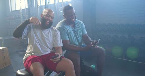 African american men laughing and checking phones while resting after gym workout