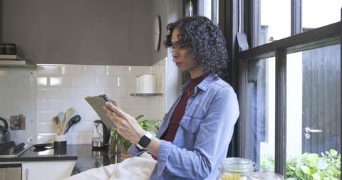 Nonbinary person on kitchen counter using tablet and checking smartwatch at sunlit window