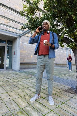 Young african american man talking on phone holding coffee on urban campus with backpack