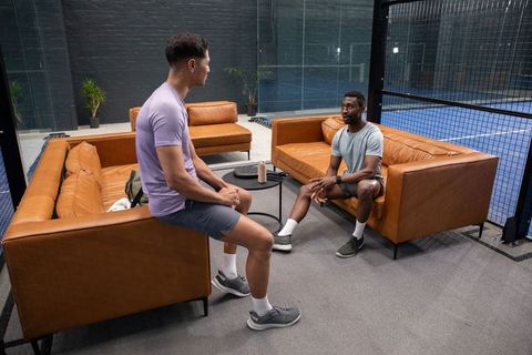 Diverse athletes chatting on leather lounge sofas beside indoor paddle tennis court