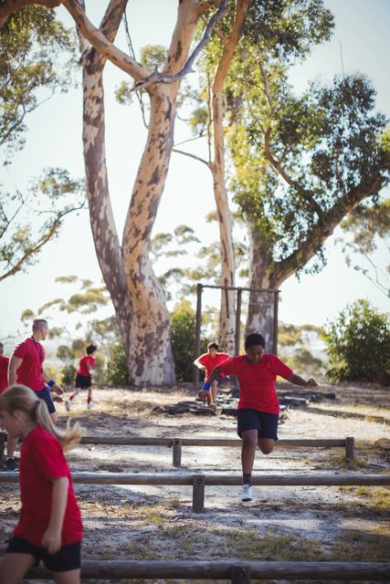 Children are actively participating in an outdoor obstacle course training session at a boot camp. They are wearing red shirts and navigating various challenges under the guidance of a trainer. This image can be used for promoting outdoor activities, fitness programs for kids, team-building exercises, and summer camps.