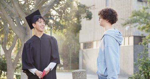 Smiling asian graduate celebrating on campus path with friend in hoodie during golden hour