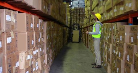 Warehouse worker inspecting inventory in high-rack storage wearing hard hat and safety vest