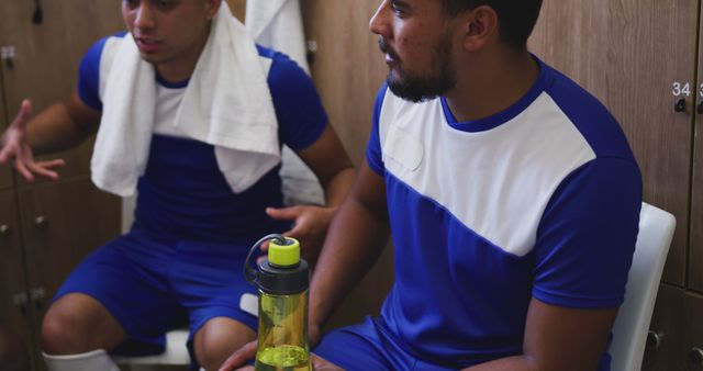 Athletes in locker room wearing blue sports jerseys and towels, hydrating while engaging in strategic discussion. Perfect for sports-related articles, fitness promotions, athletic apparel advertising, teamwork, and recovery themes.