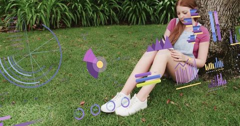 Young woman sitting under tree using smartphone with augmented data visuals on green lawn