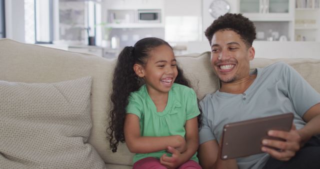 Father and daughter sharing a light moment while using a digital tablet on the couch. The warm home environment emphasizes familial bonds and modern parenting, perfect for illustrating technology use in family-friendly contexts or for advertisements promoting family interactions and digital devices.