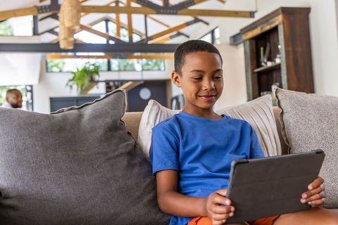 African american boy using tablet on couch, father relaxing in sunlit modern home