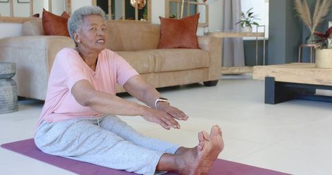 Senior african woman doing seated forward stretch on yoga mat at home wearing smartwatch