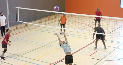 Diverse male volleyball team playing indoor gym, setter lifting colorful ball over net