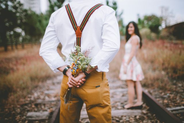 Man holding flower bouquet behind back, surprising girlfriend standing in front on railroad tracks. Ideal for romance, wedding proposals, love themes, relationship sentiments, and dating website promotions.