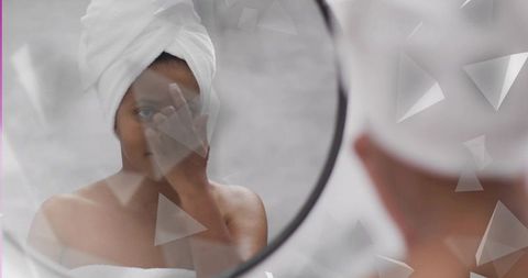 Midadult woman examining skin in mirror touching eye wearing towel turban for skincare